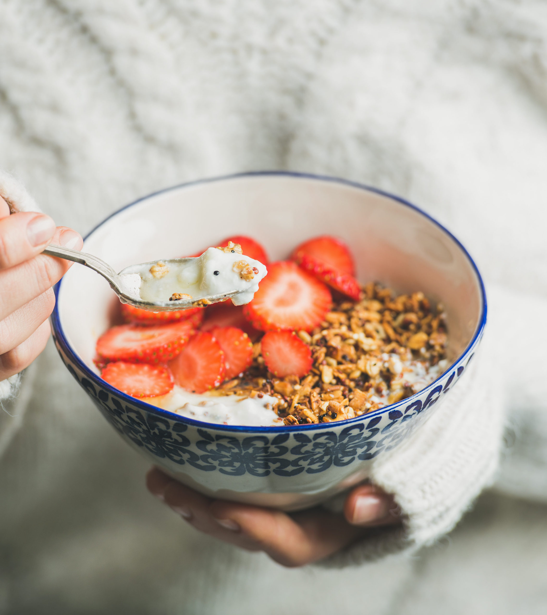 Healthy breakfast greek yogurt, gluten free granola, and strawberry bowl in hands of woman wearing white loose knitted woolen sweater